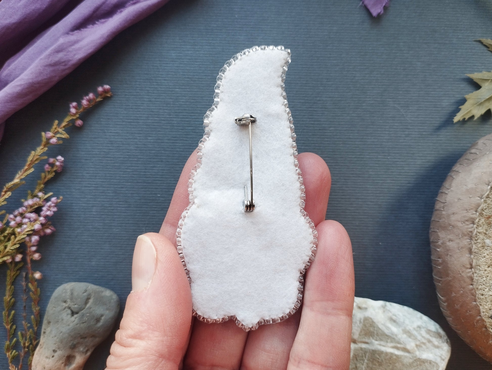A hand holding a white fabric pin with a silver pin attached to it, surrounded by various natural elements such as flowers, rocks, and leaves.