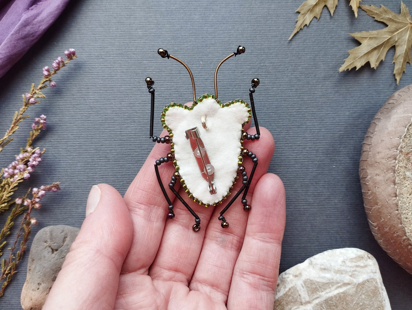 A hand holding a small, intricately designed insect-like object, which appears to be a beaded or embroidered piece.