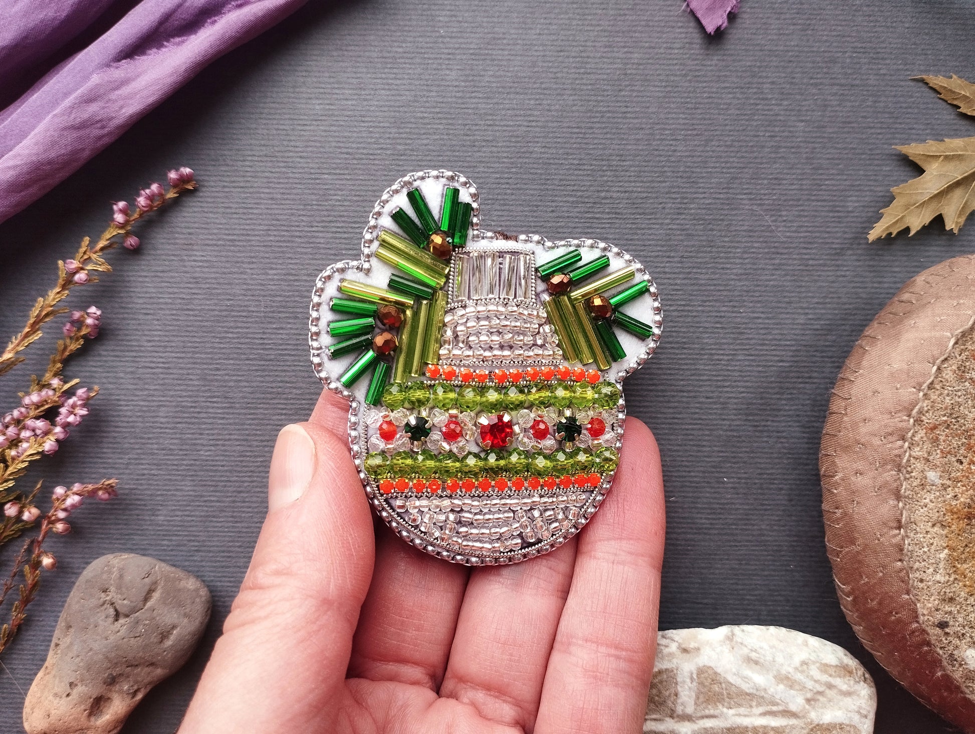 A hand holding a decorative beaded object with a floral pattern, surrounded by various natural elements such as leaves, flowers, and rocks.