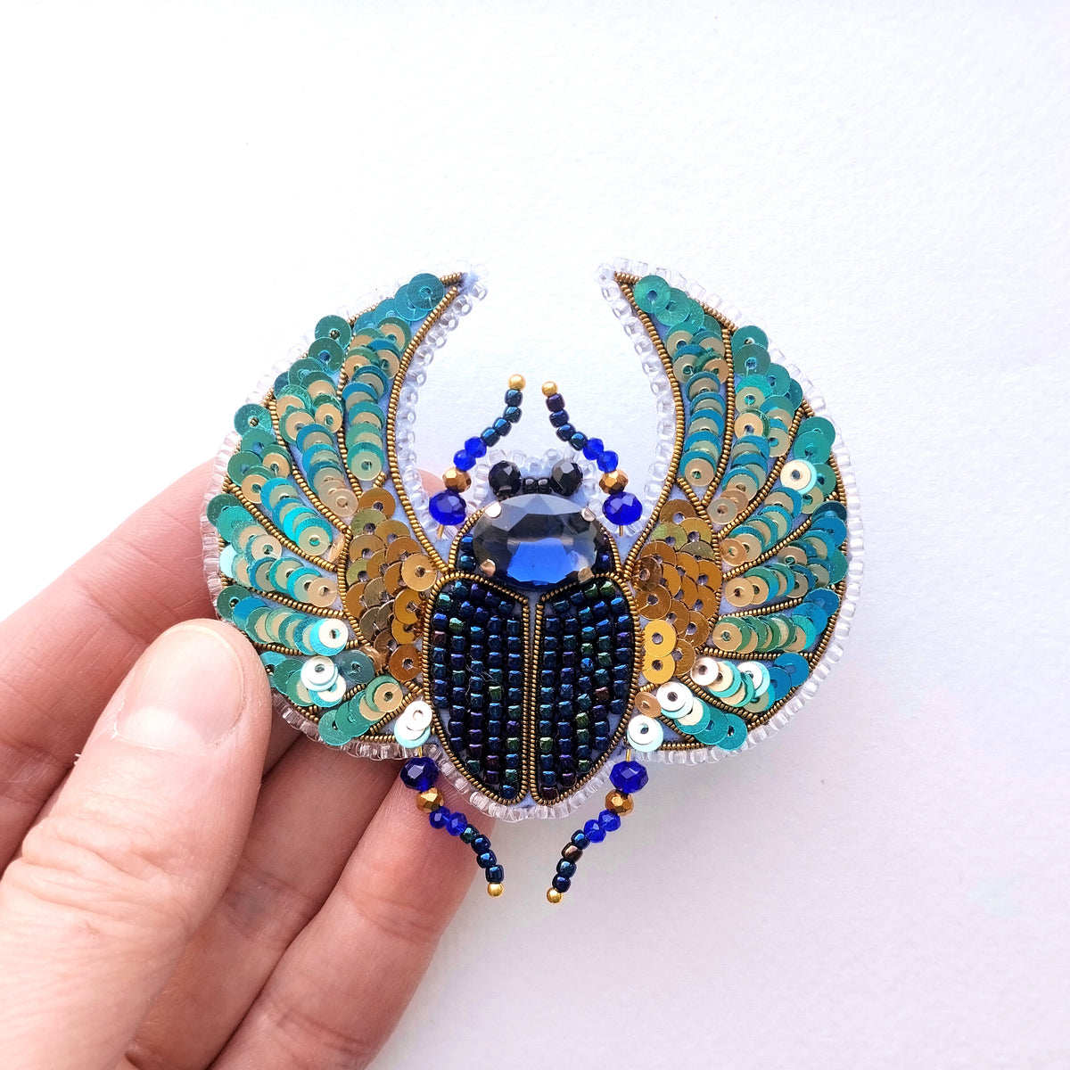 A hand is holding a decorative brooch featuring a detailed embroidered design of a blue and gold beetle.