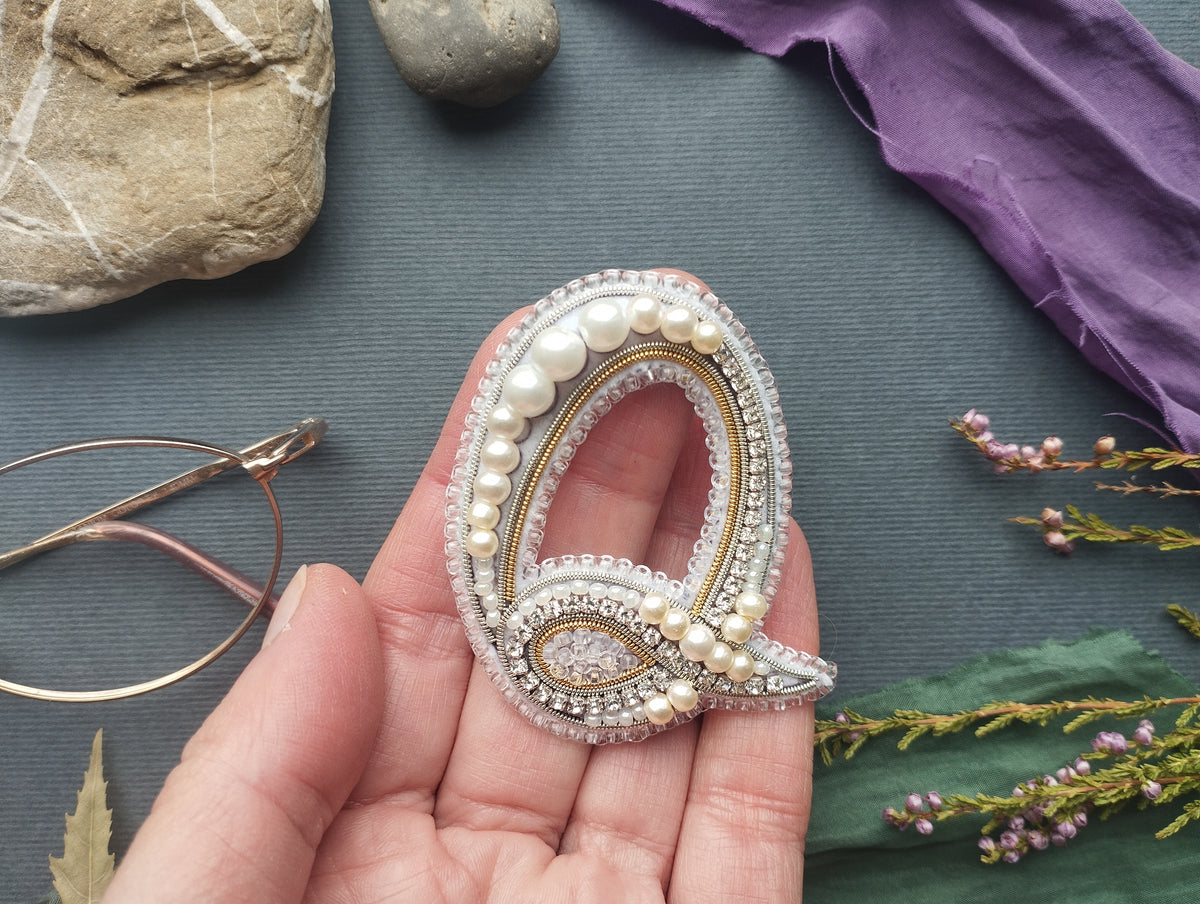 A hand holding a decorative letter "Q" made of pearls and jewels, surrounded by various objects such as a rock, a leaf, and a pair of glasses.