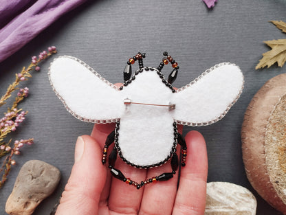 A hand holding a beaded, hand-sewn insect-shaped brooch against a gray background with various natural elements, such as leaves and rocks.
