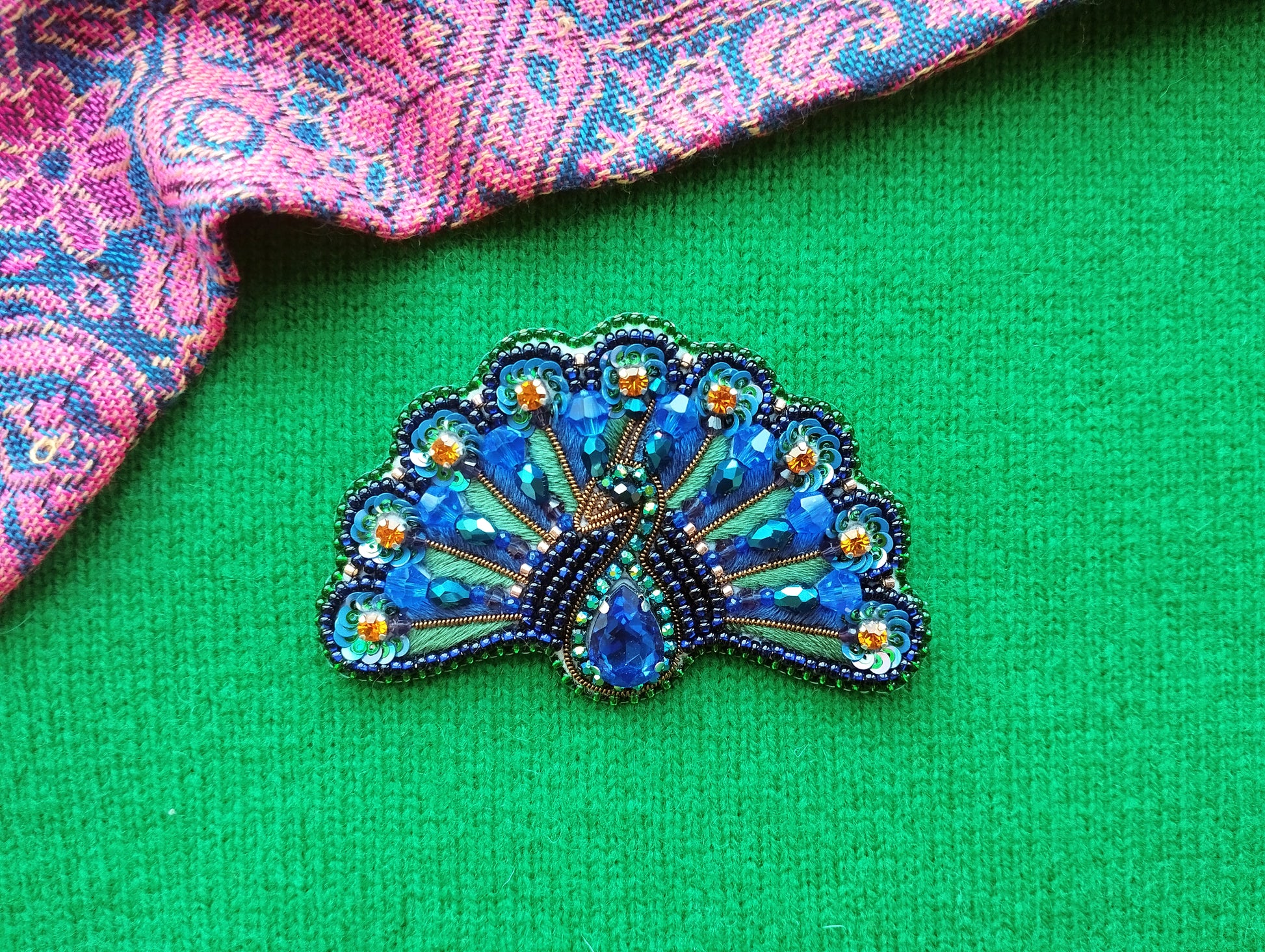 A decorative peacock brooch with blue and green beads and a gold center is displayed on a green fabric background.