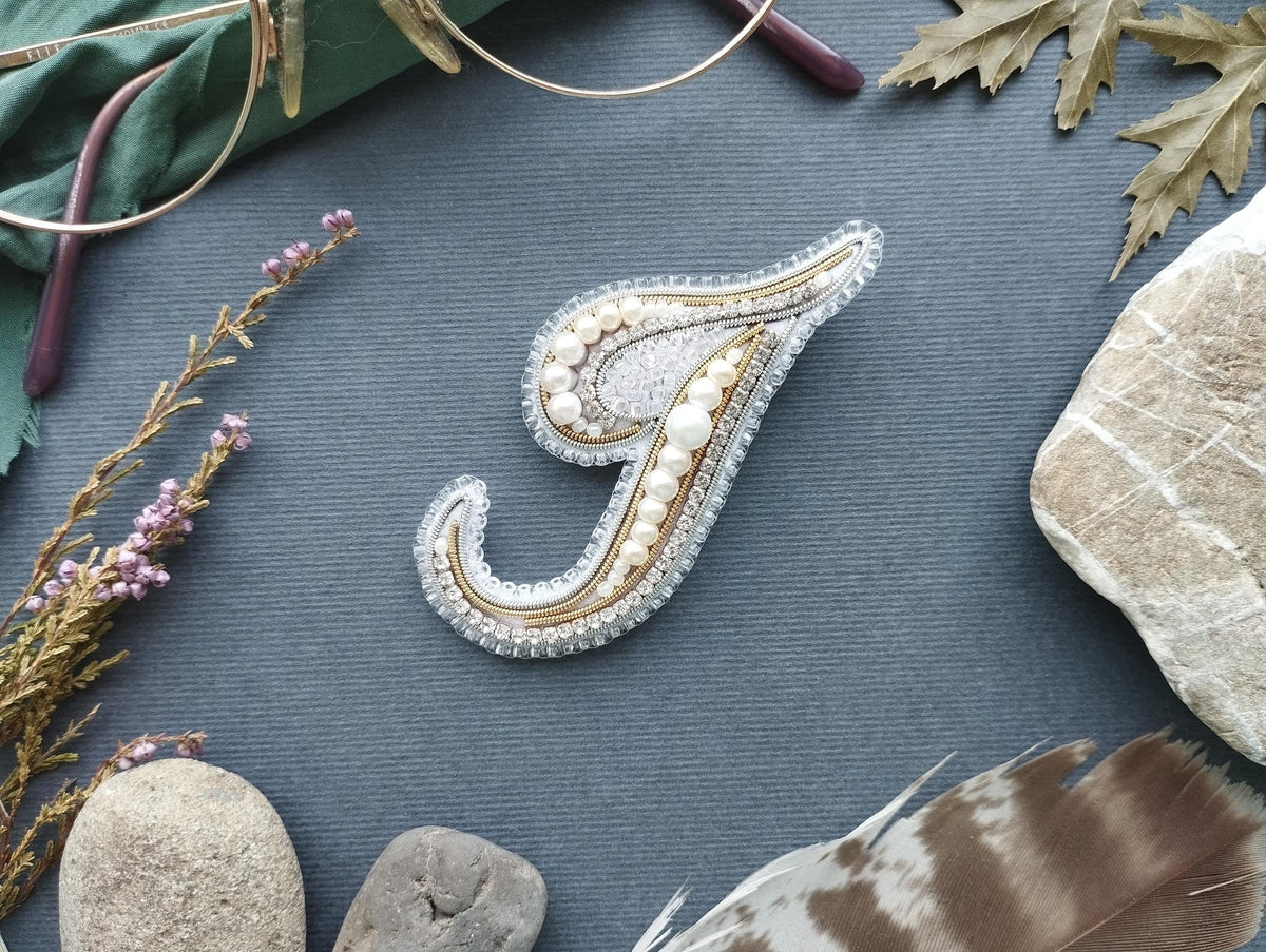 a close-up of a decorative letter 'J' made of beads and pearls, surrounded by various natural elements such as feathers, rocks, and leaves.