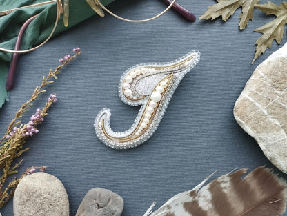 a close-up of a decorative letter 'J' made of beads and pearls, surrounded by various natural elements such as feathers, rocks, and leaves.