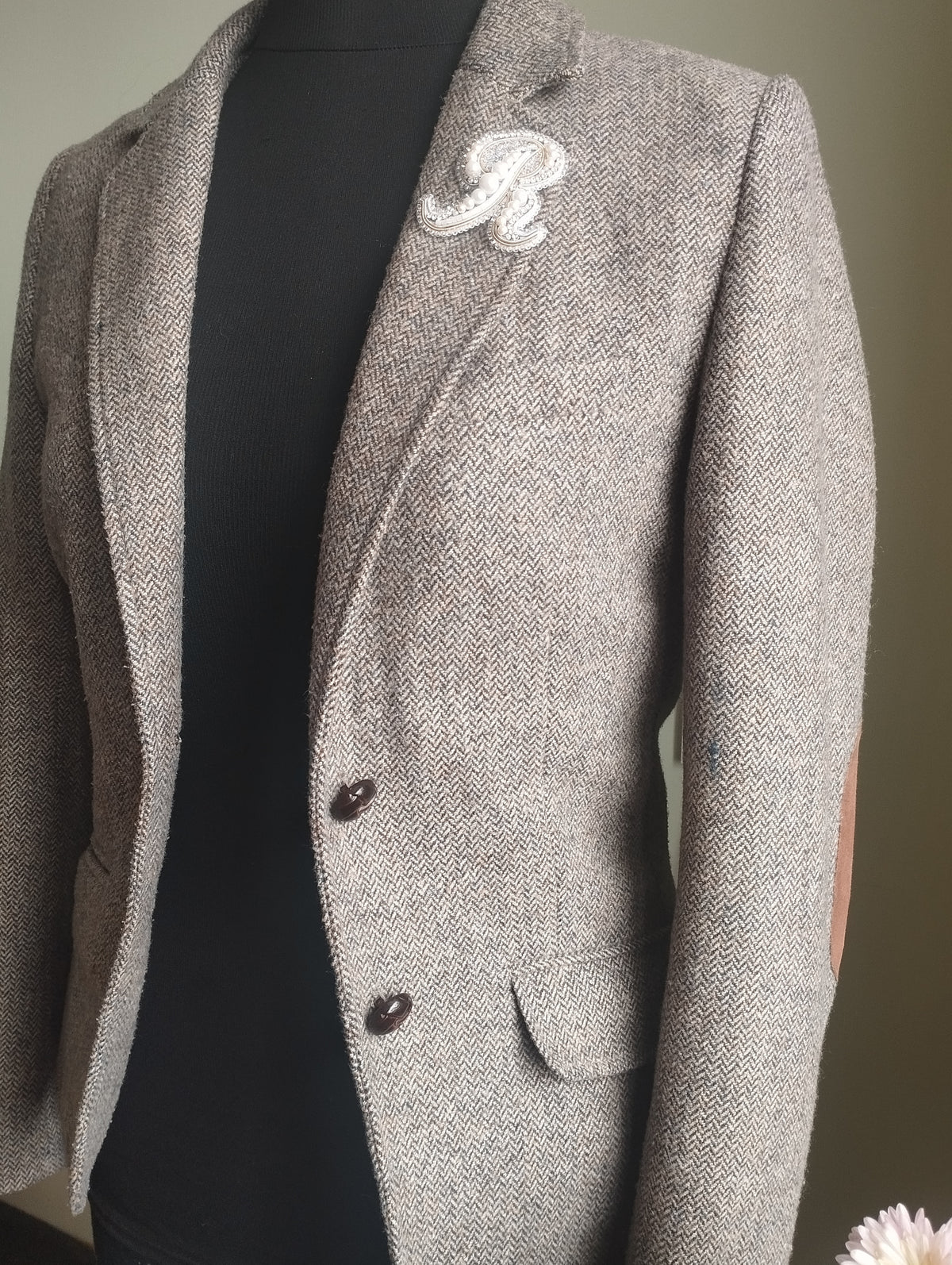 A gray tweed blazer with a white embroidered letter "R" on the lapel, displayed on a mannequin.