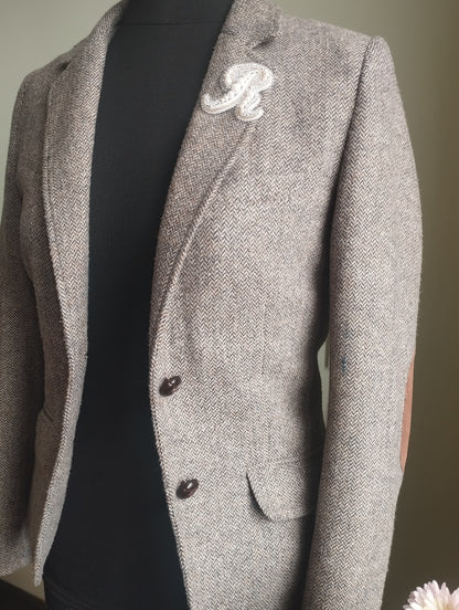 A gray tweed blazer with a white embroidered letter "R" on the lapel, displayed on a mannequin.