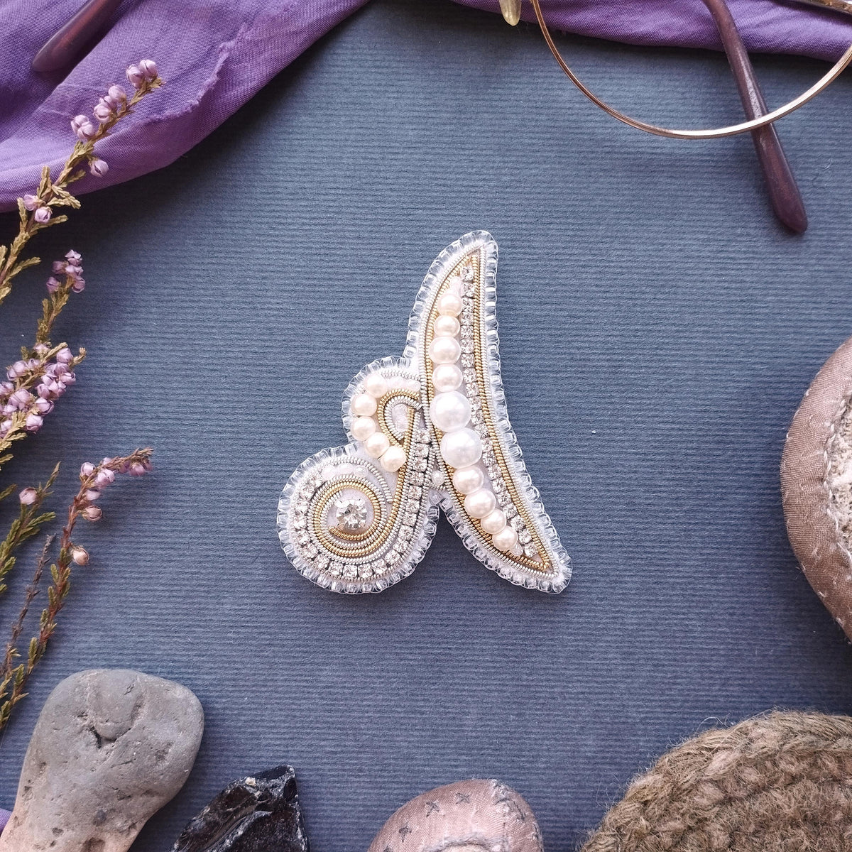 a close-up view of a decorative piece, possibly a brooch or a pin, with intricate beading and pearl details. The piece is placed on a textured surface, surrounded by various other objects such as a pair of glasses, a plant, and a small rock.
