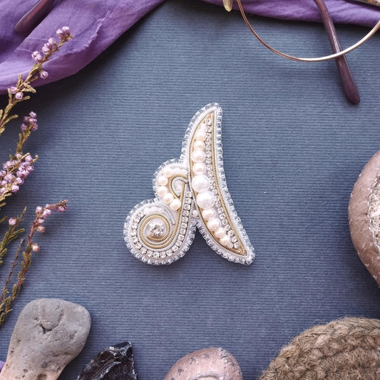 a close-up view of a decorative piece, possibly a brooch or a pin, with intricate beading and pearl details. The piece is placed on a textured surface, surrounded by various other objects such as a pair of glasses, a plant, and a small rock.