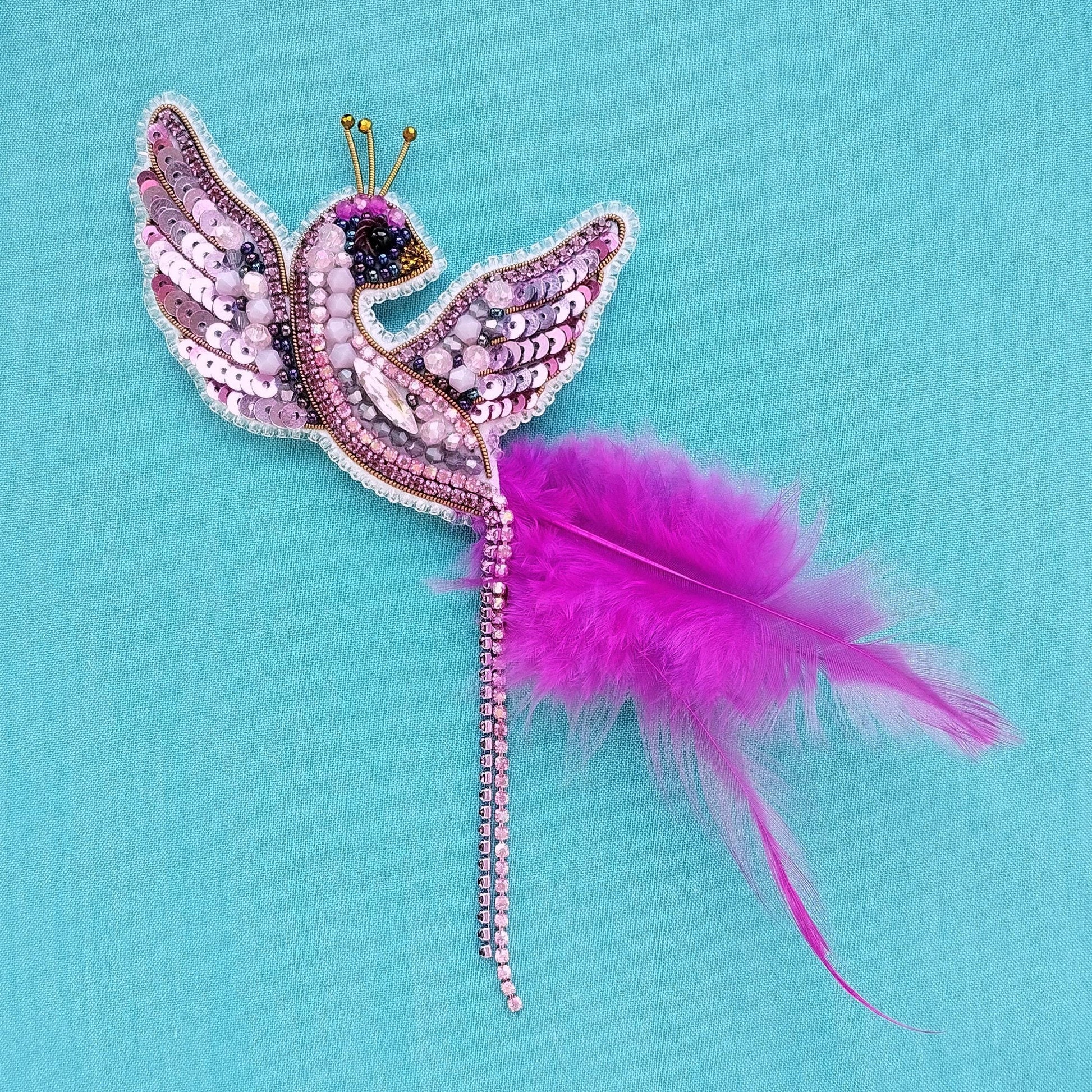 A decorative bird brooch with a pink feather and sequin wings is displayed against a blue background.