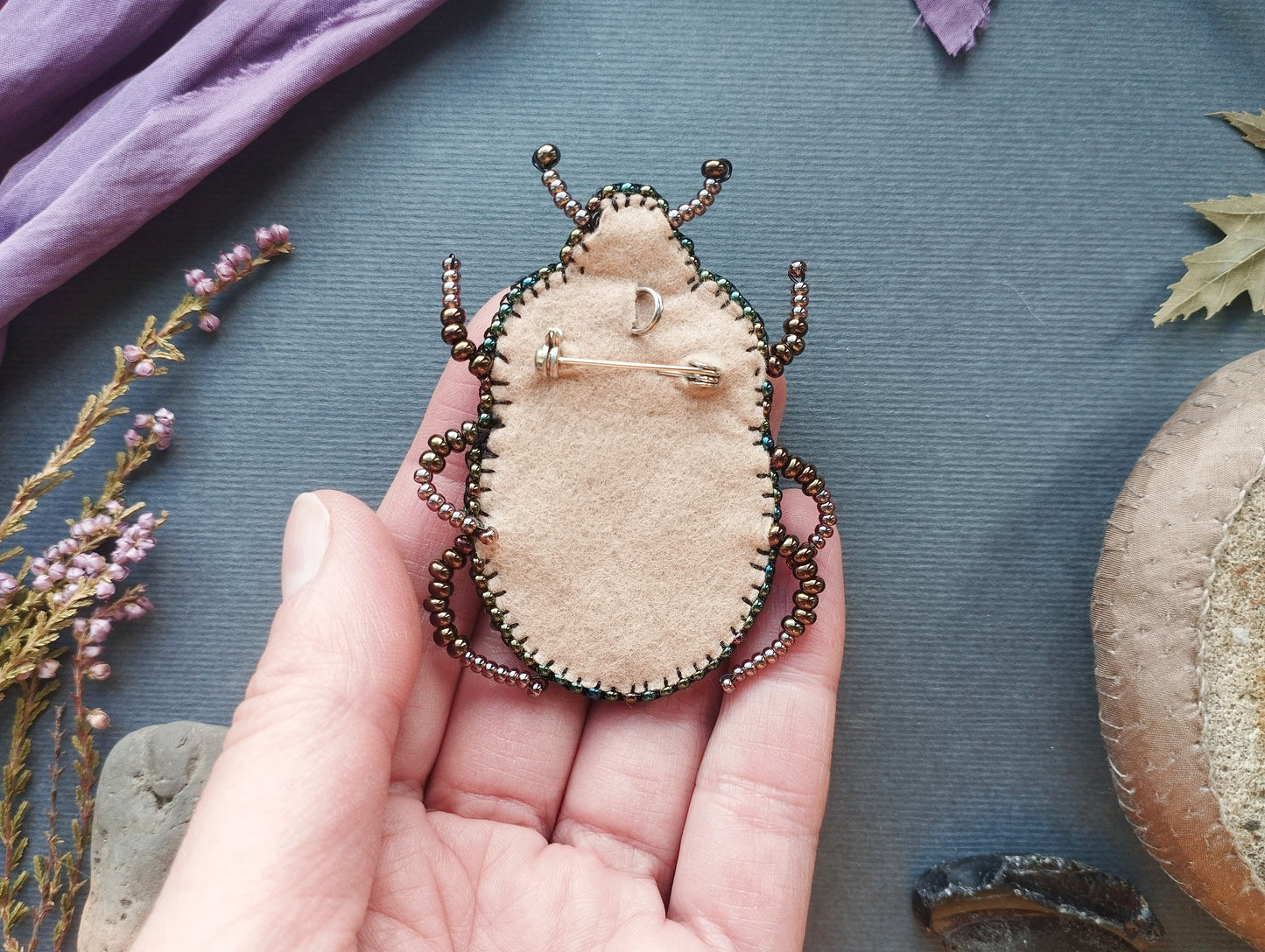 A hand holding a small, beige-colored pin-like object with a decorative design, surrounded by various natural elements such as leaves, flowers, and rocks.