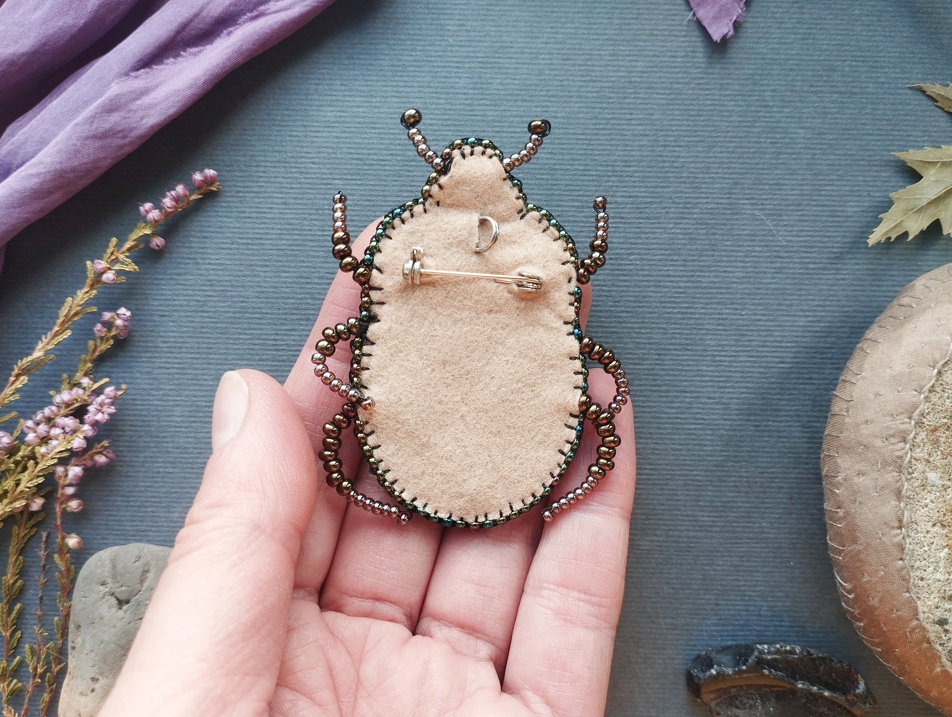 A hand holding a small, beige-colored pin-like object with a decorative design, surrounded by various natural elements such as leaves, flowers, and rocks.