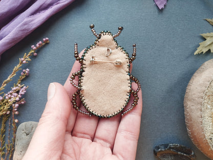 A hand holding a small, beige-colored pin-like object with a decorative design, surrounded by various natural elements such as leaves, flowers, and rocks.