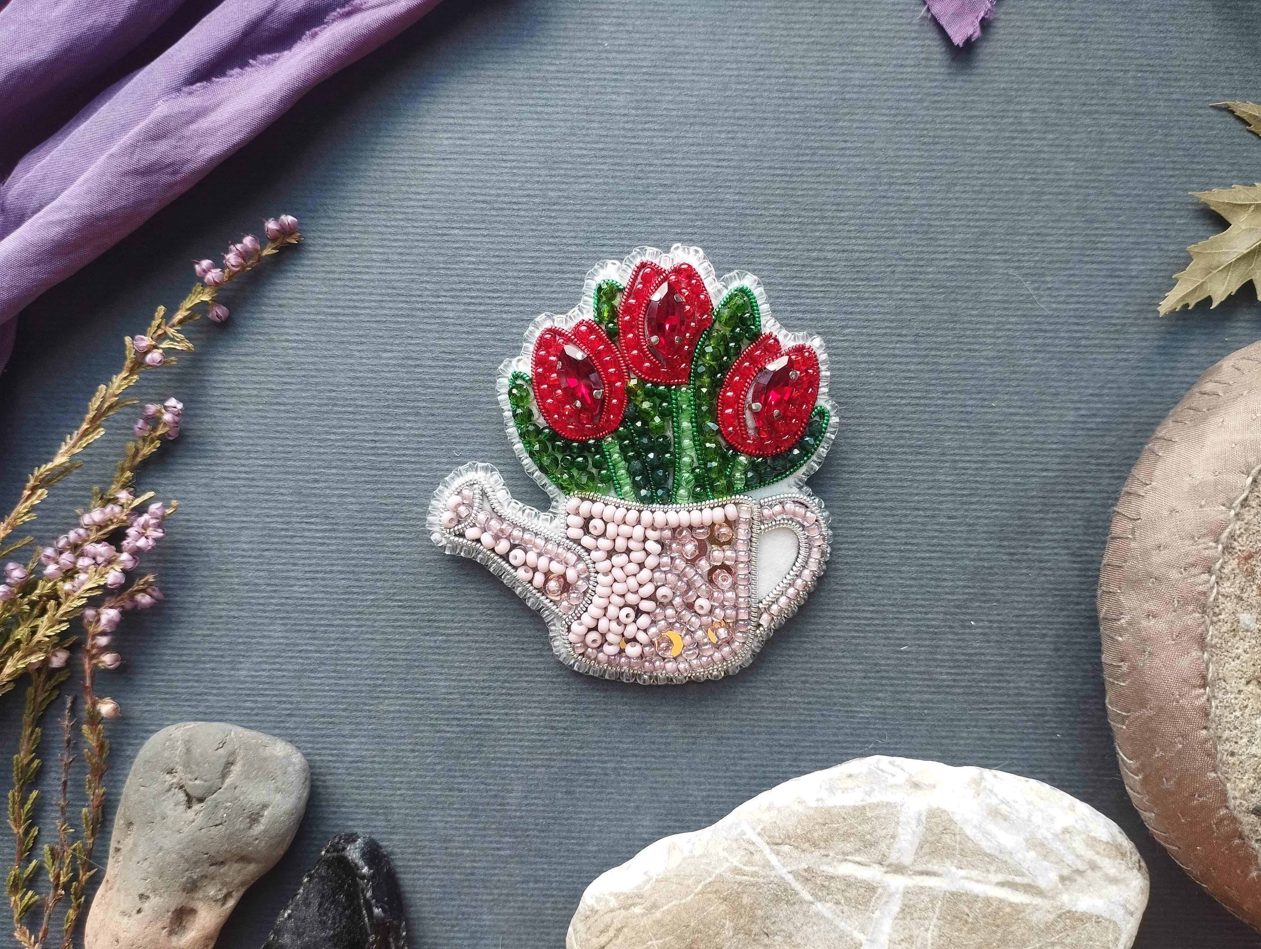 a decorative piece of art, a beaded flower in the shape of a watering can, surrounded by various natural elements such as flowers, rocks, and leaves.