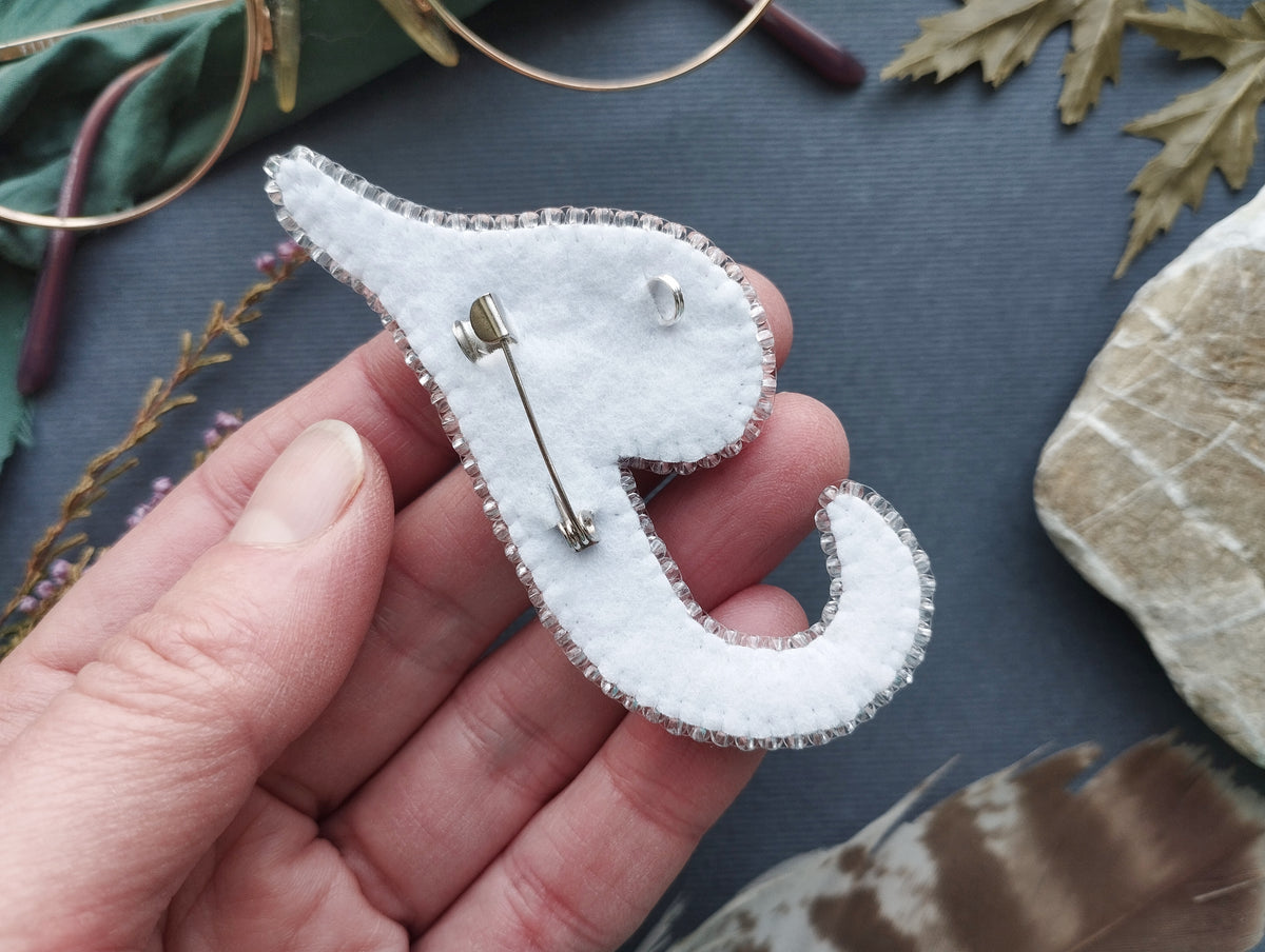 A hand holding a white felt pin with a silver pin attached to it, surrounded by various objects such as feathers and rocks.