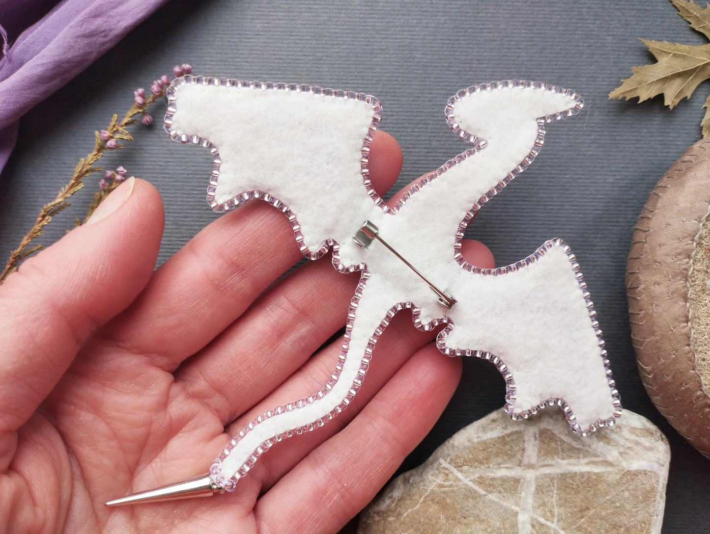 A hand holding a white felt dragon pin against a gray background with some natural elements like leaves and a rock.