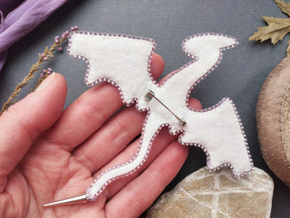 A hand holding a white felt dragon pin against a gray background with some natural elements like leaves and a rock.
