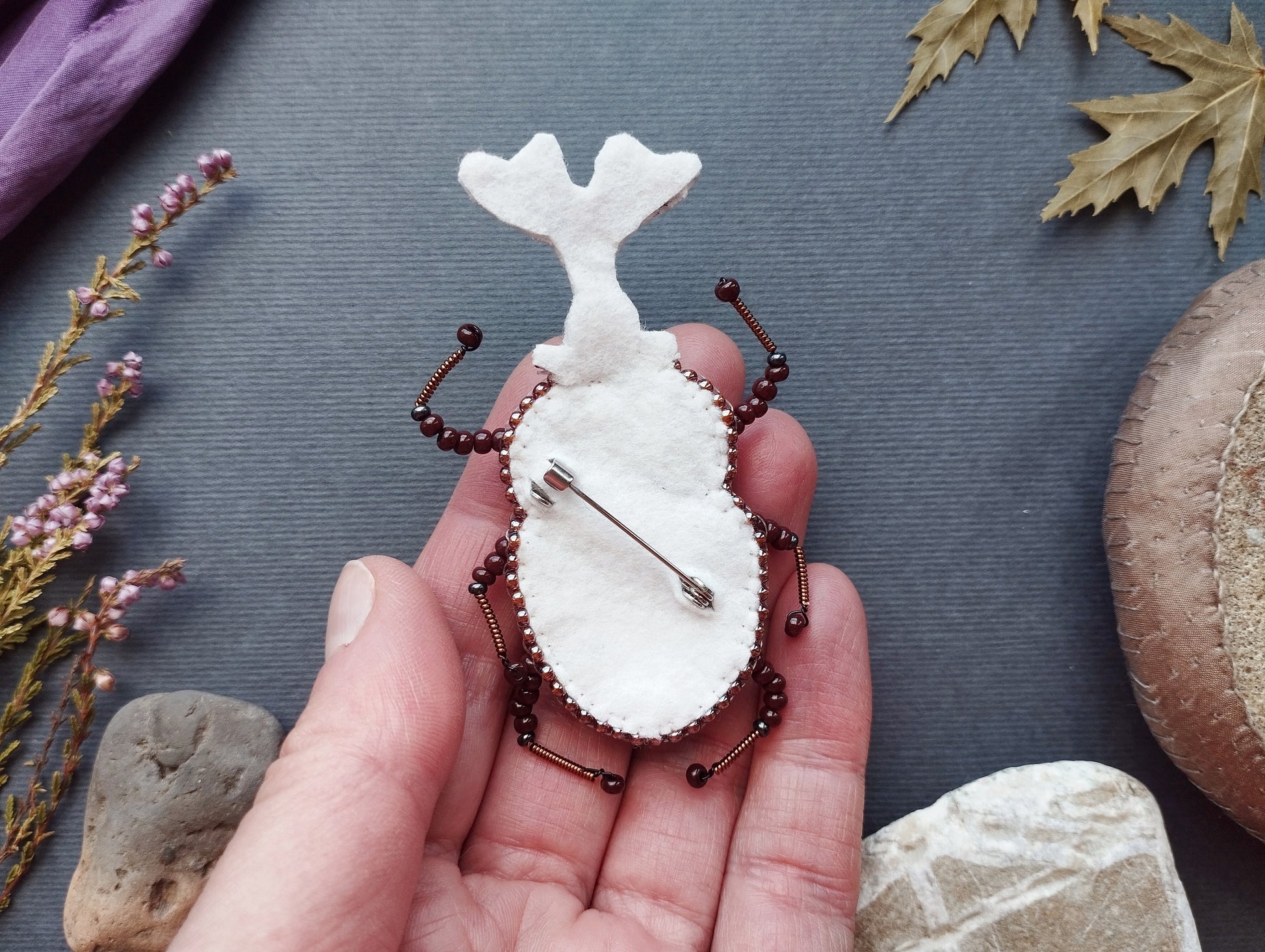 A hand holding a white, beaded insect-shaped pin against a dark background with various natural elements, such as leaves and rocks.