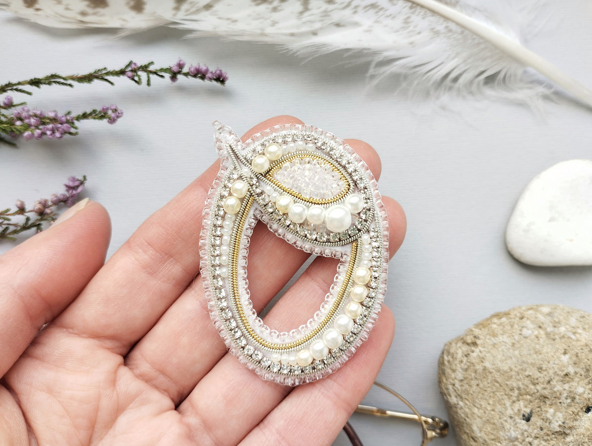 A hand holding a decorative brooch with a floral design and pearls.