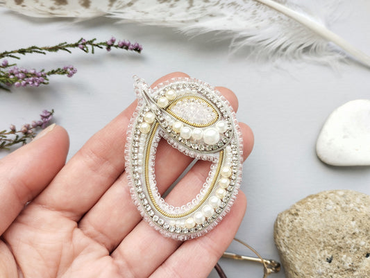 A hand holding a decorative brooch with a floral design and pearls.
