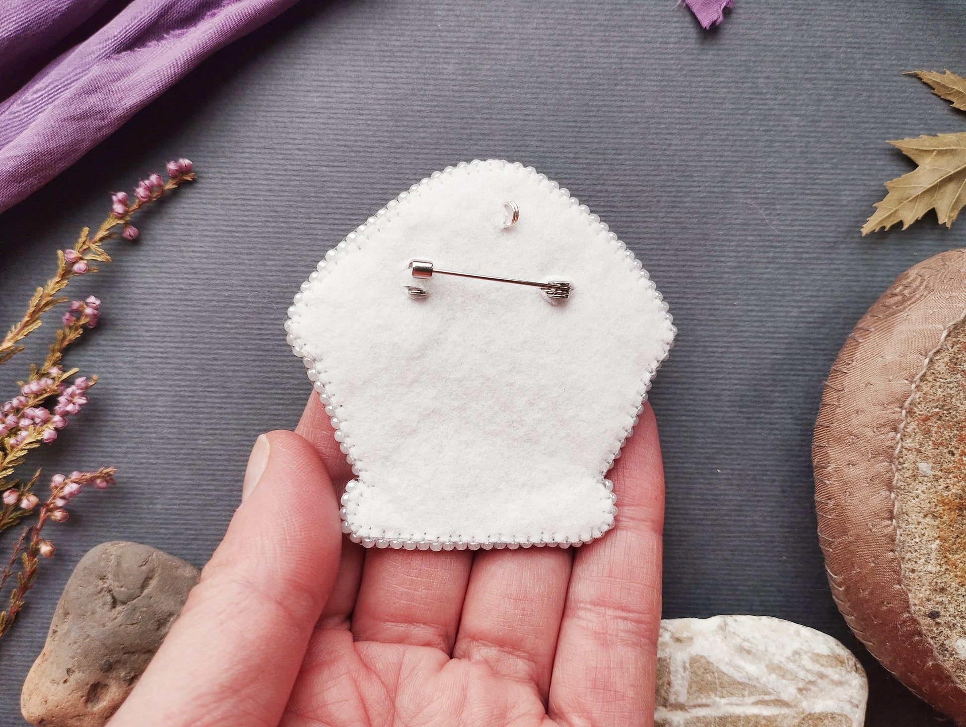 A hand holding a white felt object with a pin hole in the center, surrounded by various natural elements such as leaves, flowers, and rocks.