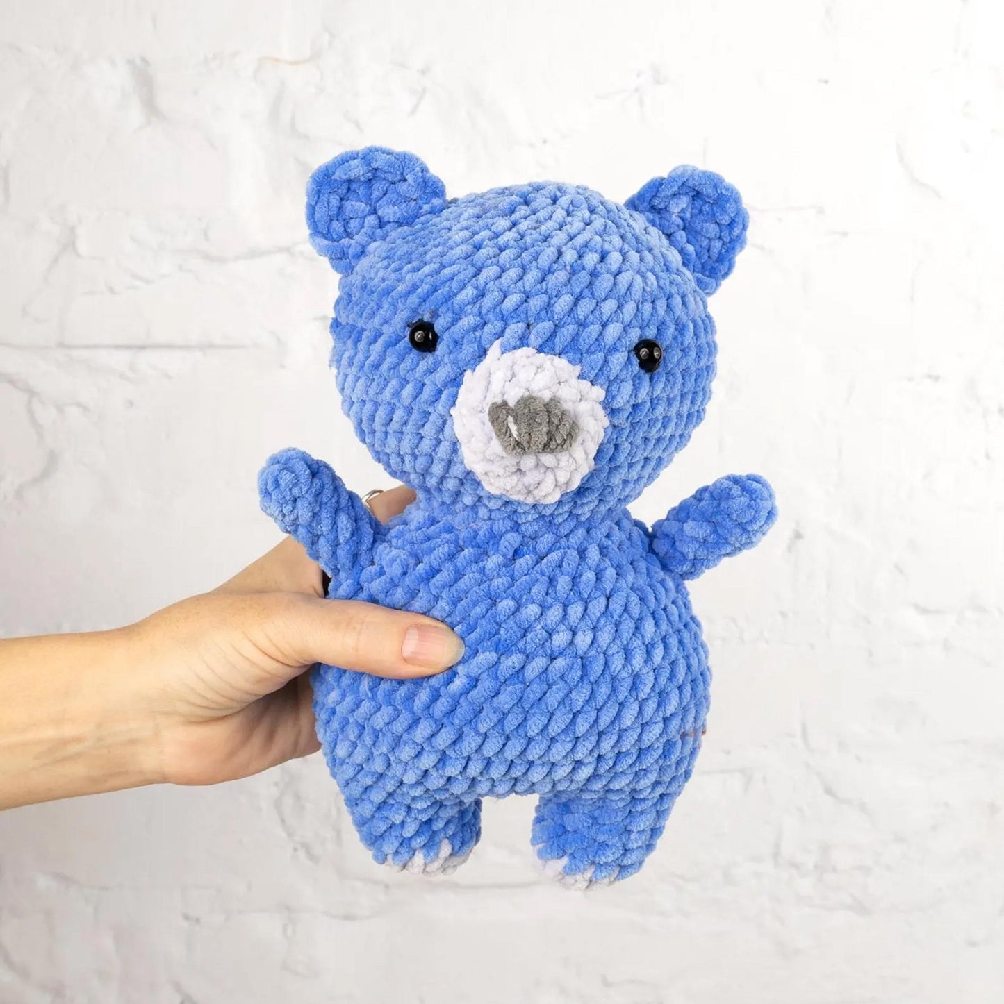 A hand is holding a blue, crocheted stuffed bear toy against a white background.