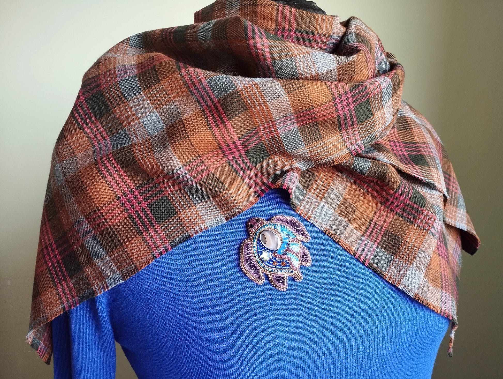 A plaid scarf is draped over a blue sweater, with a decorative brooch pinned to the sweater's back.