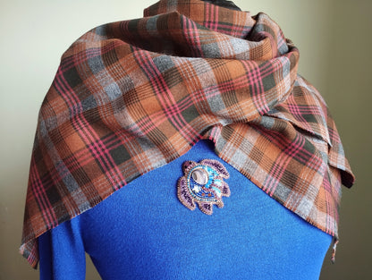 A plaid scarf is draped over a blue sweater, with a decorative brooch pinned to the sweater's back.