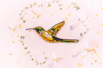 a vibrant, hand-embroidered hummingbird with a yellow body, green wings, and a black beak, surrounded by delicate white flowers and green leaves.