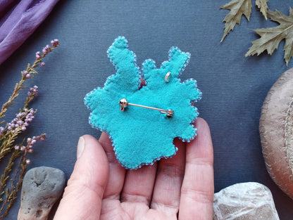 A hand holding a blue felt object with a pin attached to it, surrounded by various natural elements such as leaves, flowers, and rocks.
