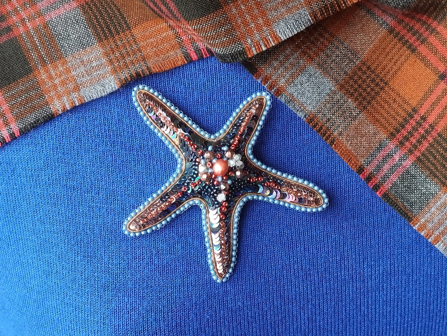A decorative starfish brooch with a blue and red color scheme, adorned with beads and pearls, is displayed against a blue fabric background.