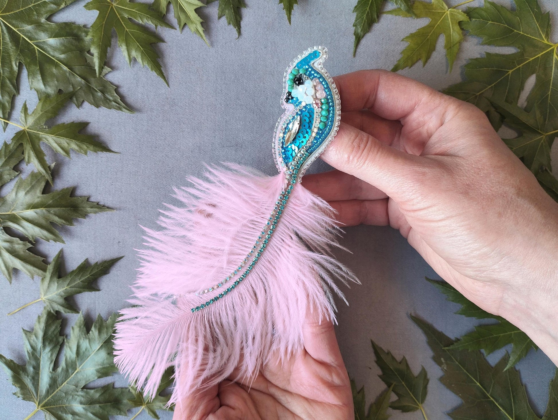A person's hand holding a decorative feather with a blue and green beaded design, surrounded by green leaves.
