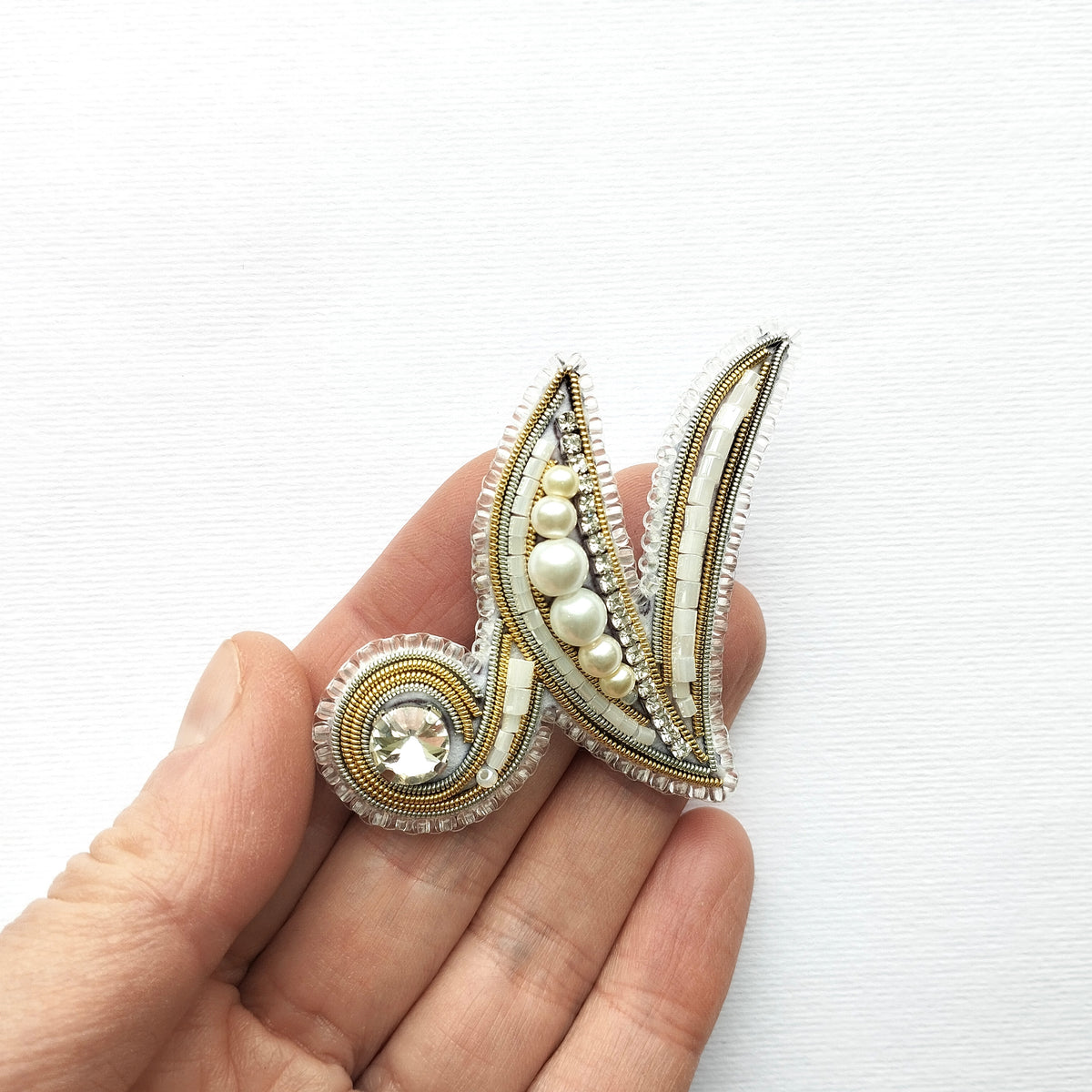 A hand holding a decorative piece of jewelry, which appears to be a brooch or pin. The piece is adorned with various embellishments, including pearls and a gemstone, giving it an elegant and sophisticated appearance.