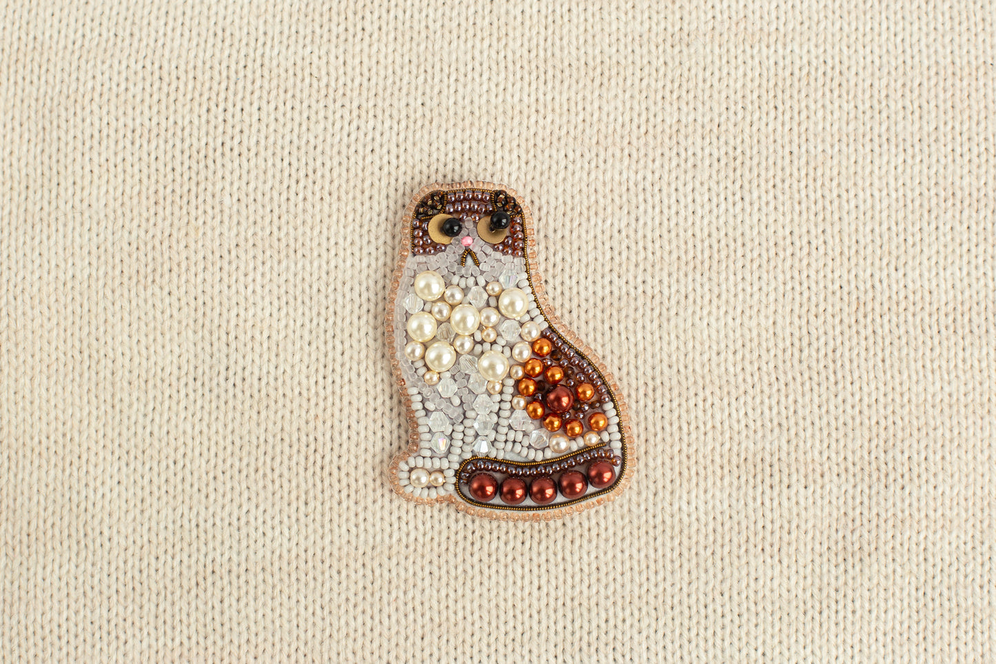 A decorative cat brooch made of beads and pearls is displayed against a beige background.