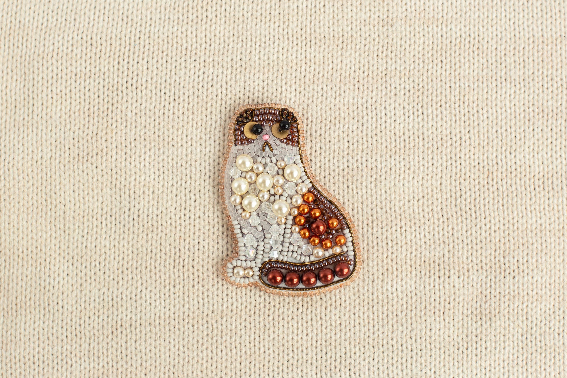 A decorative cat brooch made of beads and pearls is displayed against a beige background.