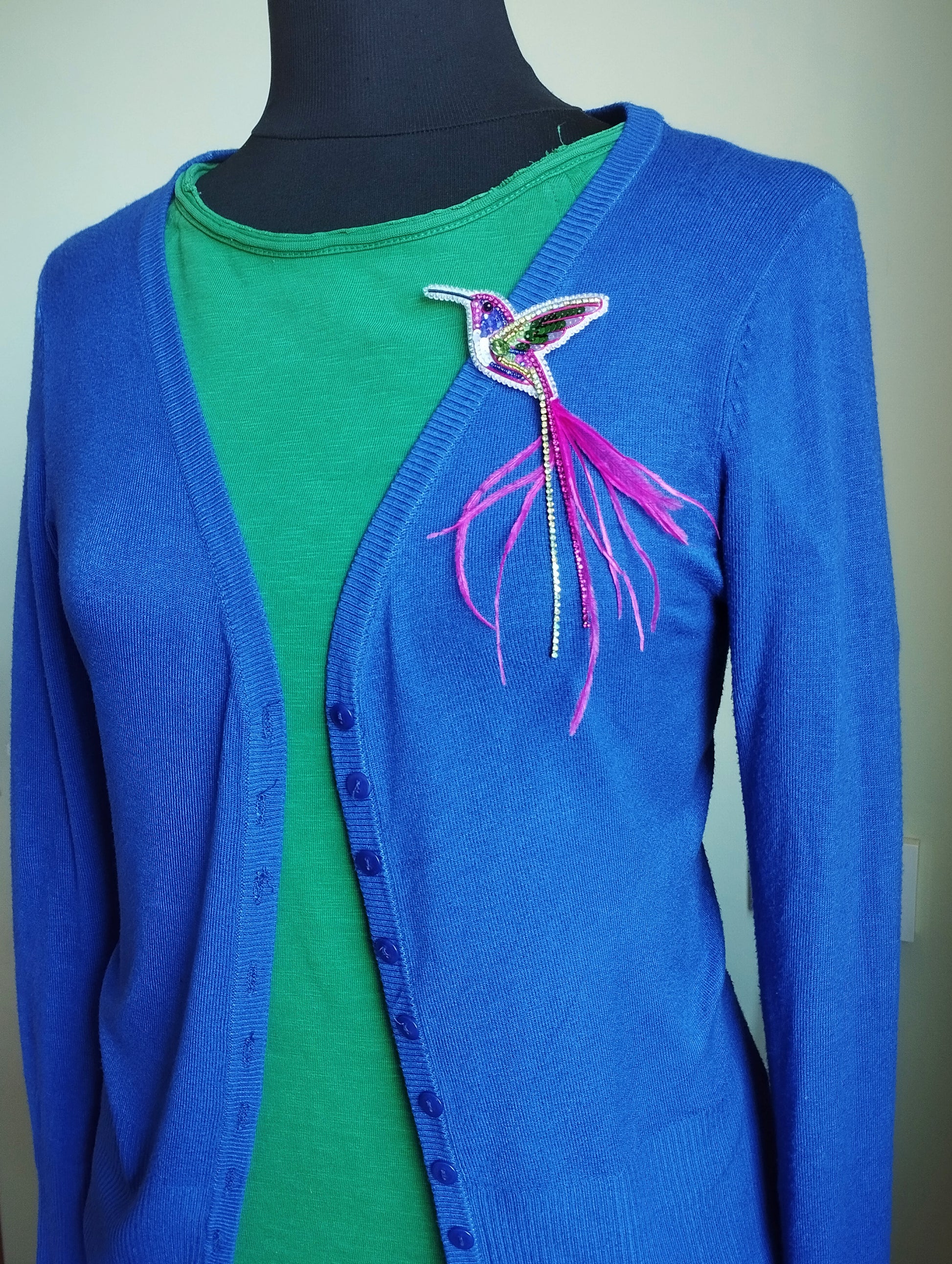 A mannequin wearing a blue cardigan with a green shirt underneath, and a colorful bird brooch on the left side of the chest.