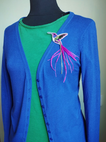 A mannequin wearing a blue cardigan with a green shirt underneath, and a colorful bird brooch on the left side of the chest.