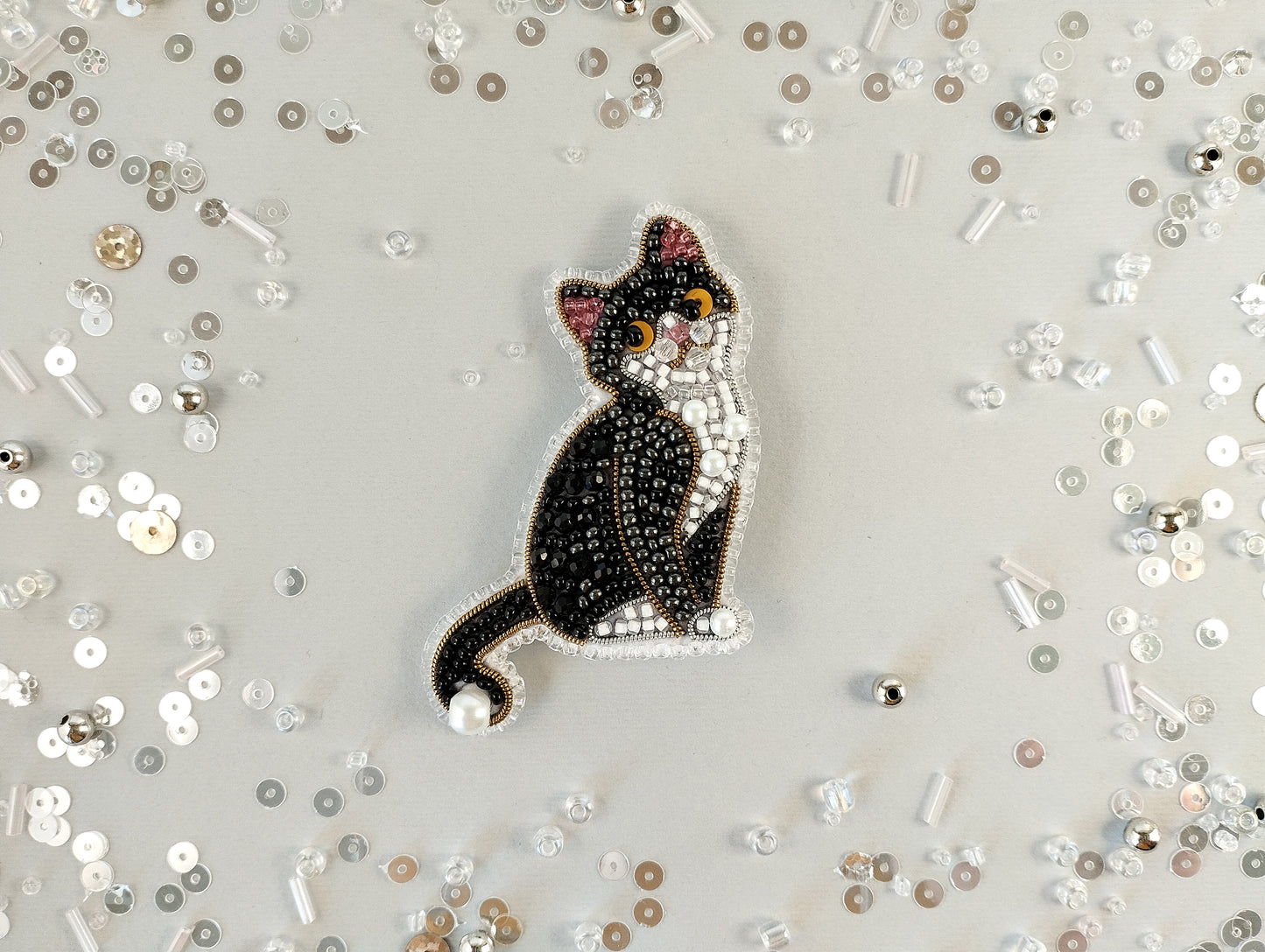 A black and white cat embroidered with sparkling beads is the central focus of the image, surrounded by scattered silver and white beads on a light background.