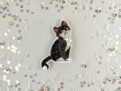 A black and white cat embroidered with sparkling beads is the central focus of the image, surrounded by scattered silver and white beads on a light background.