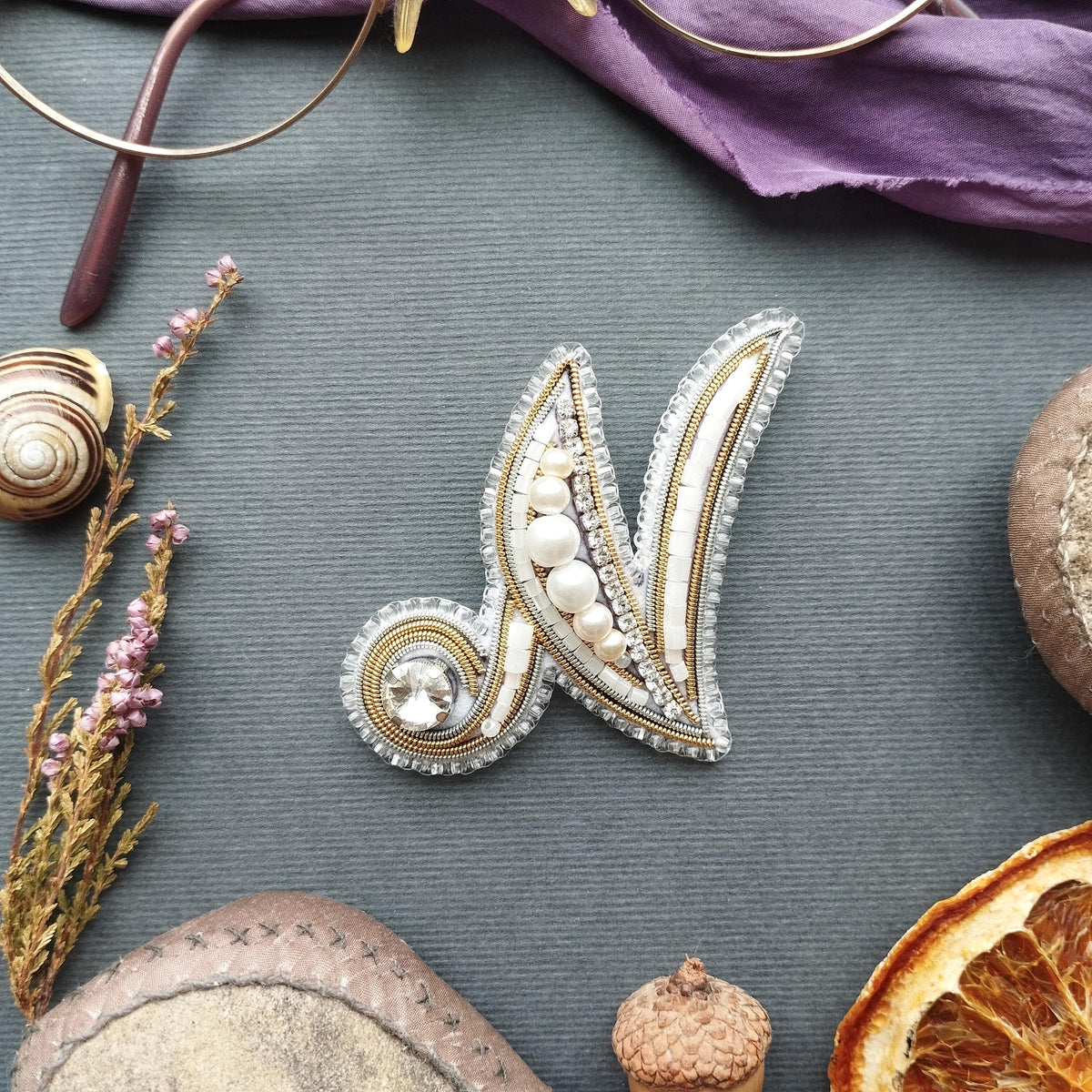 a close-up view of a decorative letter 'N' made of silver and white beads, placed on a gray surface. Surrounding the 'N' are various objects, including a pair of glasses, a seashell, a piece of fruit, and a small acorn.