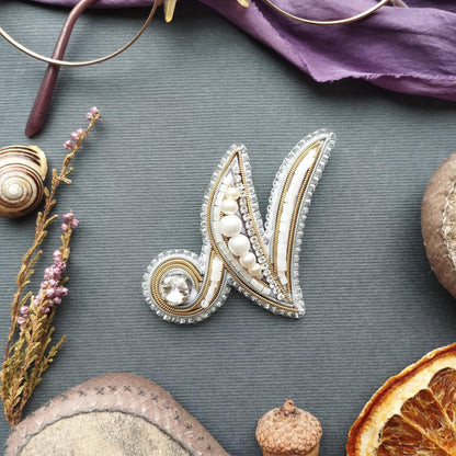 a close-up view of a decorative letter 'N' made of silver and white beads, placed on a gray surface. Surrounding the 'N' are various objects, including a pair of glasses, a seashell, a piece of fruit, and a small acorn.