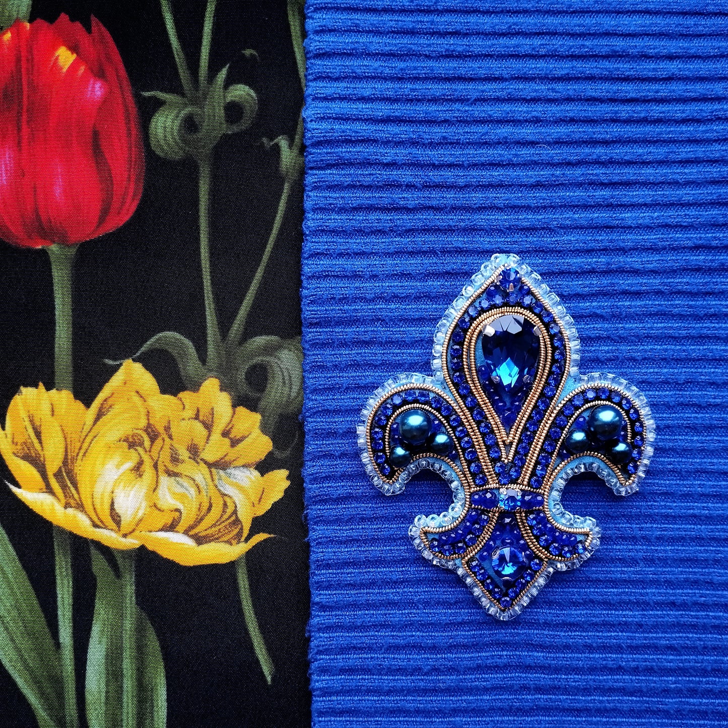 a close-up of a blue fabric with a decorative blue and gold brooch in the center, and a red tulip flower on the left side.