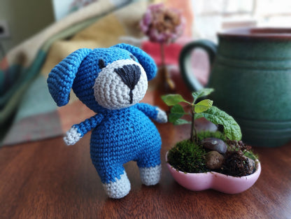 a crocheted blue teddy bear next to a plant