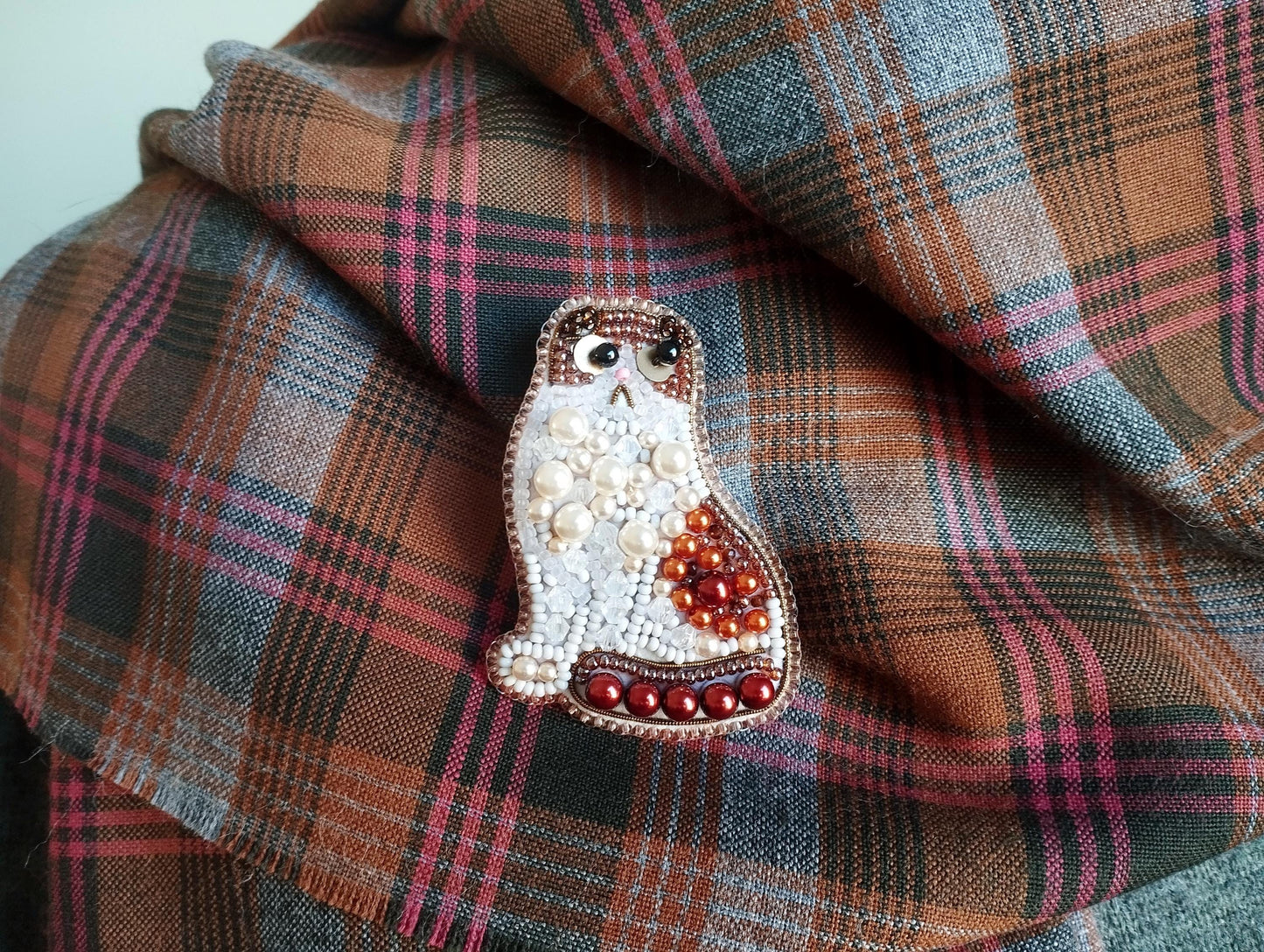 A decorative cat brooch made of beads and pearls is displayed on a plaid fabric background.