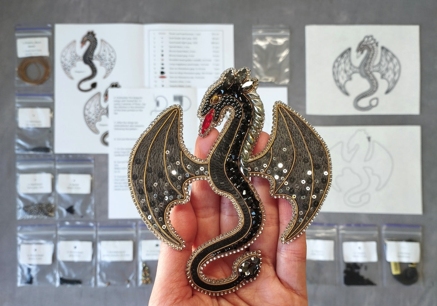 A hand is holding a decorative dragon pin, which is adorned with sequins and has a detailed design. The background includes various materials and tools, suggesting that the image might be a collage or a workspace for creating such items.