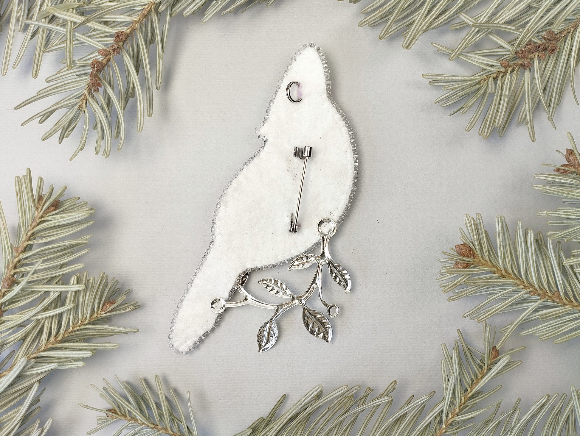 a white bird-shaped brooch with silver leaf branches attached to it, surrounded by pine needles.