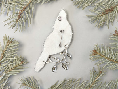 a white bird-shaped brooch with silver leaf branches attached to it, surrounded by pine needles.