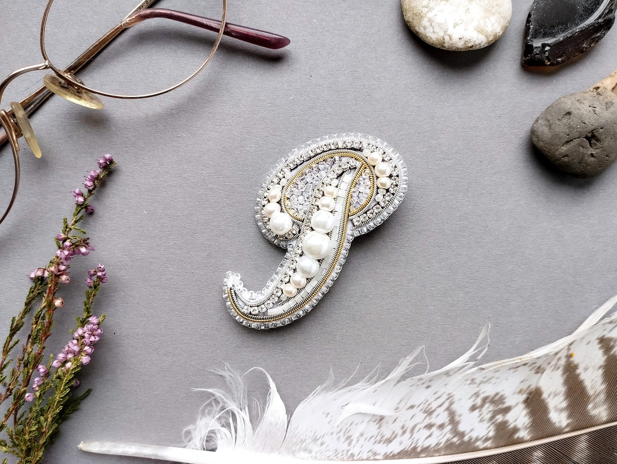 a close-up view of a decorative monogrammed letter "P" adorned with pearls and jewels, surrounded by various accessories such as glasses, feathers, and stones.