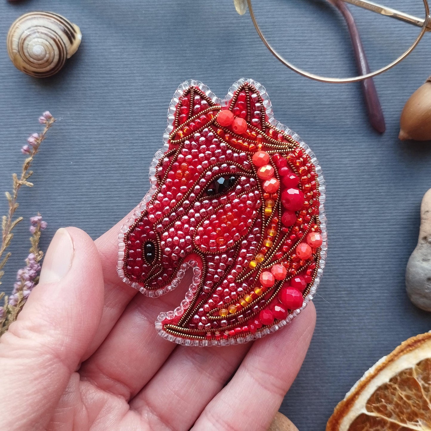 A hand holding a red and gold beaded embroidered piece resembling a horse's head, surrounded by various objects such as a snail, glasses, and dried flowers.