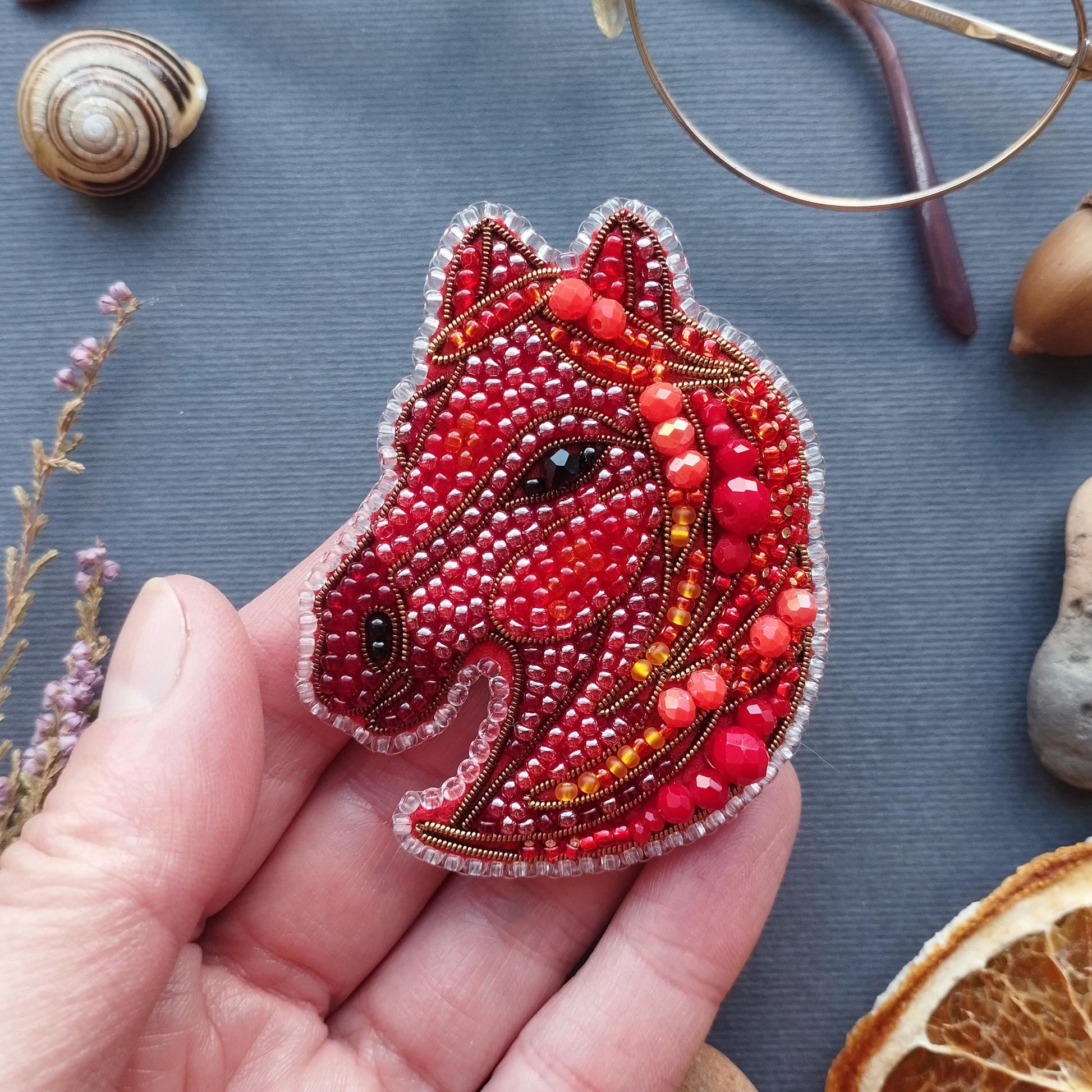 A hand holding a red and gold beaded embroidered piece resembling a horse's head, surrounded by various objects such as a snail, glasses, and dried flowers.